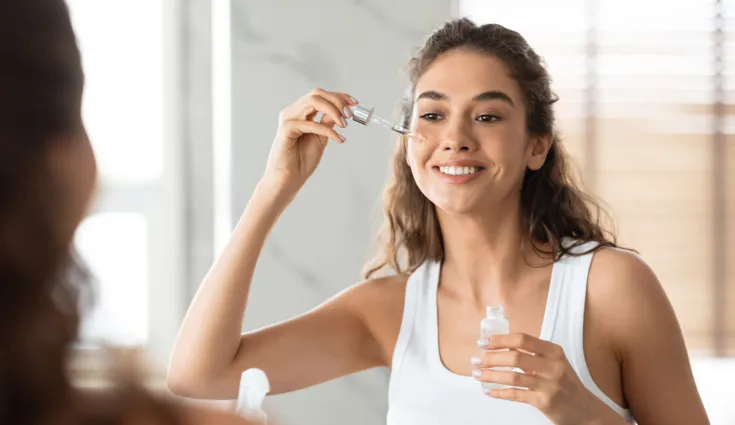 A smiling woman applies serum to her face with a dropper in a mirror.