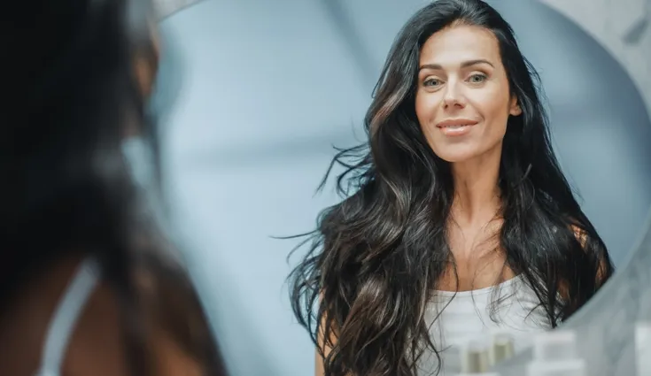 A woman with long dark hair and glowing skin smiles in a mirror.