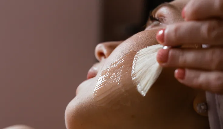 A facial brush sweeps across a woman's face, applying a clear solution to her cheek.