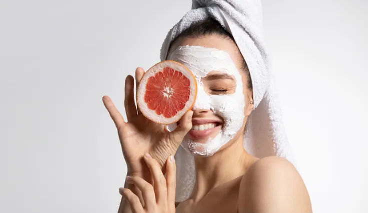 A smiling woman wears a white face mask while holding a grapefruit slice up to her eye.