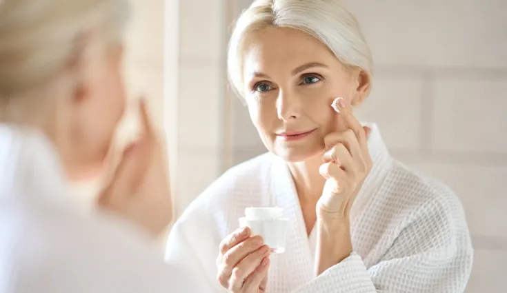 Mature women applying moisturizer to her face while looking in the mirror